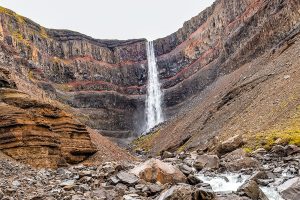 Visiting Hengifoss: Iceland’s Iconic Red-Strata Waterfall A tall, narrow waterfall plunging down a deep gorge framed by dark basalt cliff walls with prominent horizontal stripes of red clay and sedimentary rock in East Iceland.