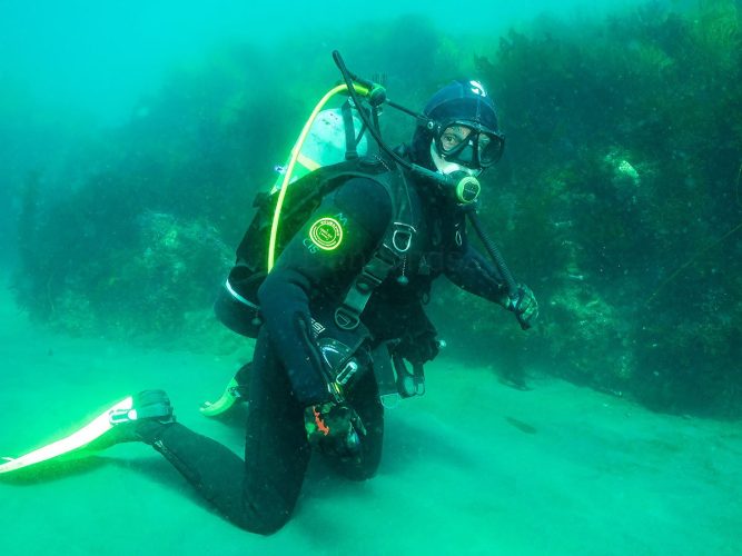 Exploring the underwater world: Dan Lindsay geared up for a cold-water dive alongside a reef structure, showcasing the technical equipment required to comfortably navigate and capture these vibrant marine environments. Underwater portrait of a male scuba diver, Dan Lindsay, kneeling on a sandy ocean floor next to a dark reef wall covered in marine growth. He is wearing full cold-water diving gear, including a black thick wetsuit, hood, gloves, mask, and a Scubapro buoyancy compensator device (BCD). A silver scuba tank with bright yellow accents is mounted on his back, and his bright yellow fins rest on the sand against a clear turquoise water background.