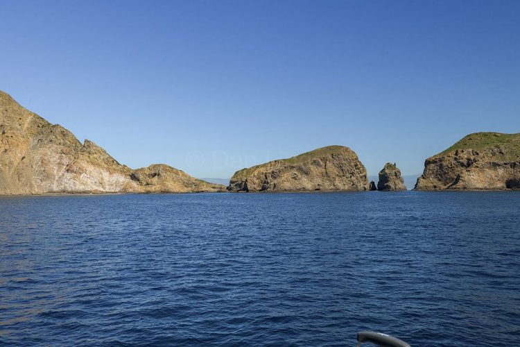 Looking across the deep blue waters toward the rugged volcanic cliffs and sea caves of the Channel Islands. A wide-angle landscape view from the ocean looking toward a series of rugged, steep rocky islands with sparse green vegetation on top under a clear blue sky.